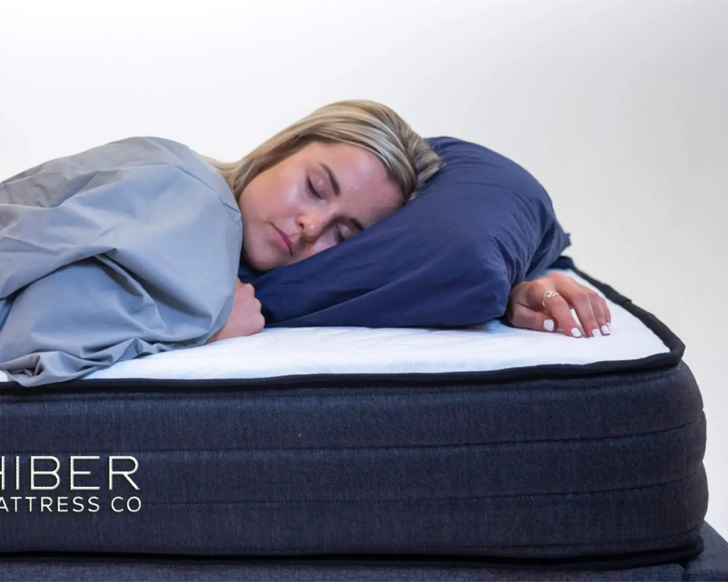 Person sleeping on a mattress with a pillow, wearing a blanket, on a white background. Hiber Mattress Co branding visible.